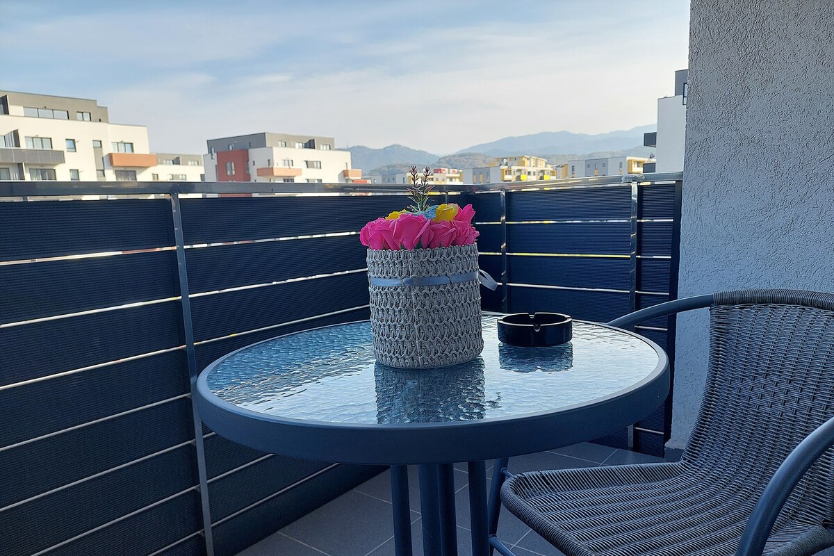 A round table with a woven vase of flowers, a cigarette pack, and a chair on a balcony with a view of buildings and mountains.