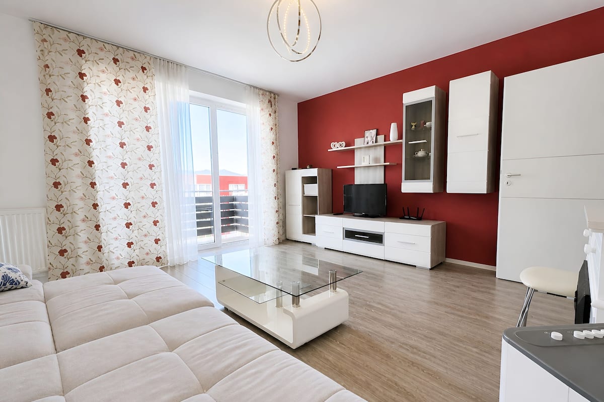 A modern living room with a sofa, a glass coffee table, a TV, and a red accent wall.