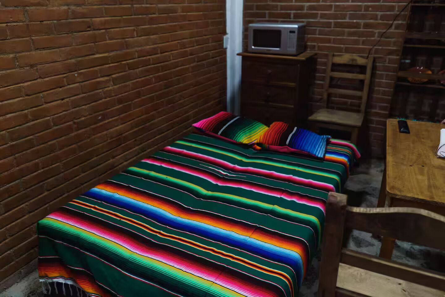 A small, narrow room with a brick wall, a wooden bed with colorful striped bedding, a small wooden nightstand with a microwave, and a wooden chair.