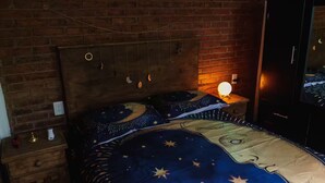 Un dormitorio con pared de ladrillo, cabecera de madera, cama con colcha de estampado de estrellas, mesita de noche con lámpara y televisor en la pared.