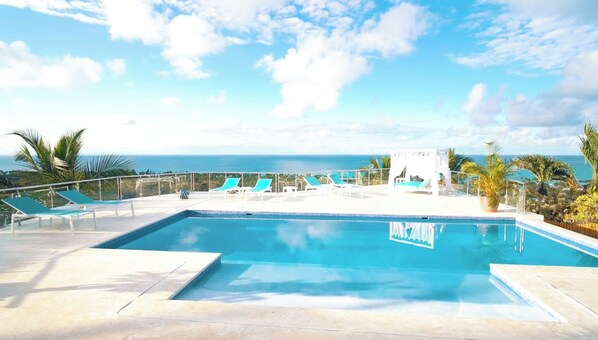 Zona de piscina con vista al océano, sillas de descanso y palmeras.