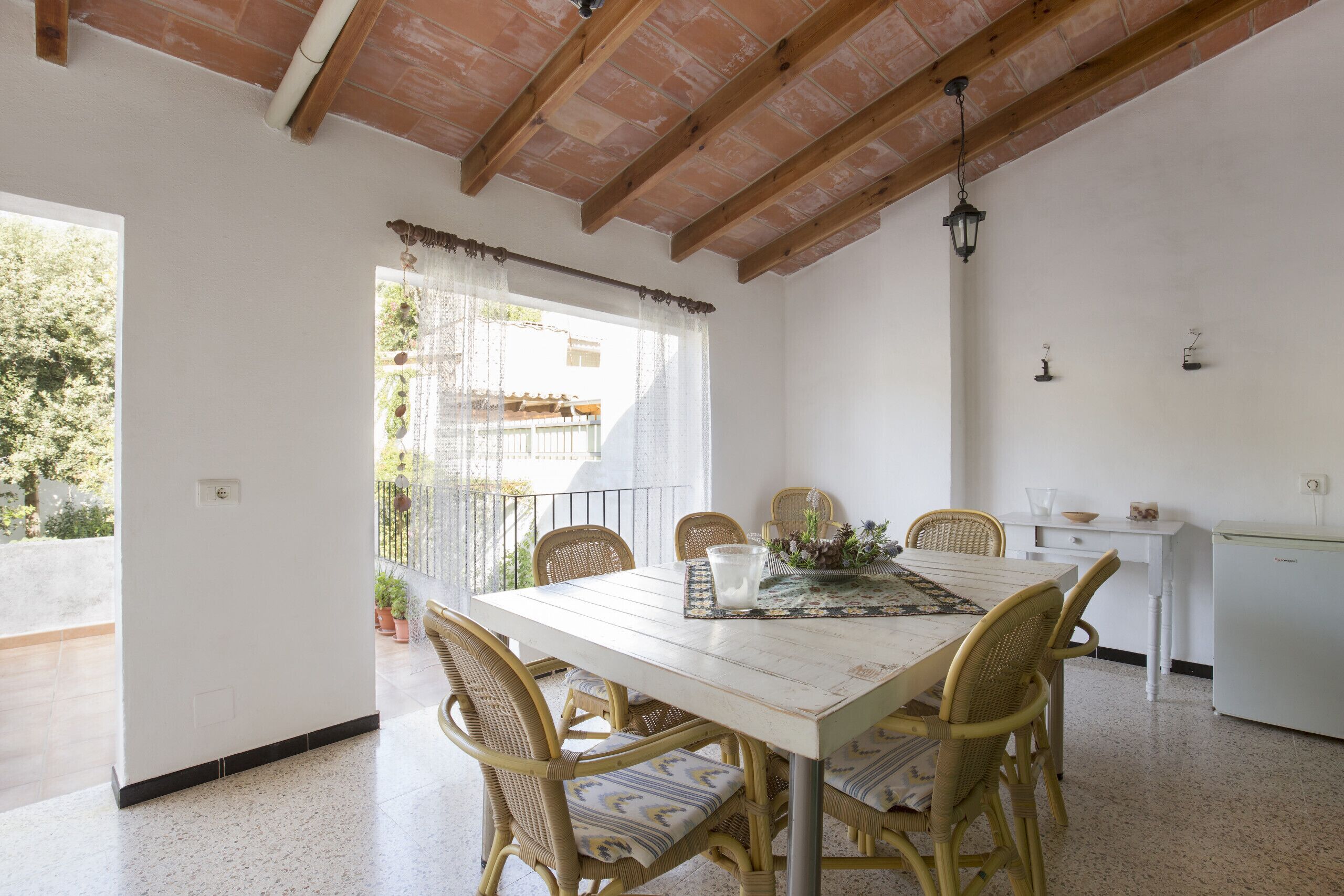 A dining area with a white table and wicker chairs, a kitchen area with a white refrigerator, and a balcony with a railing and potted plants.