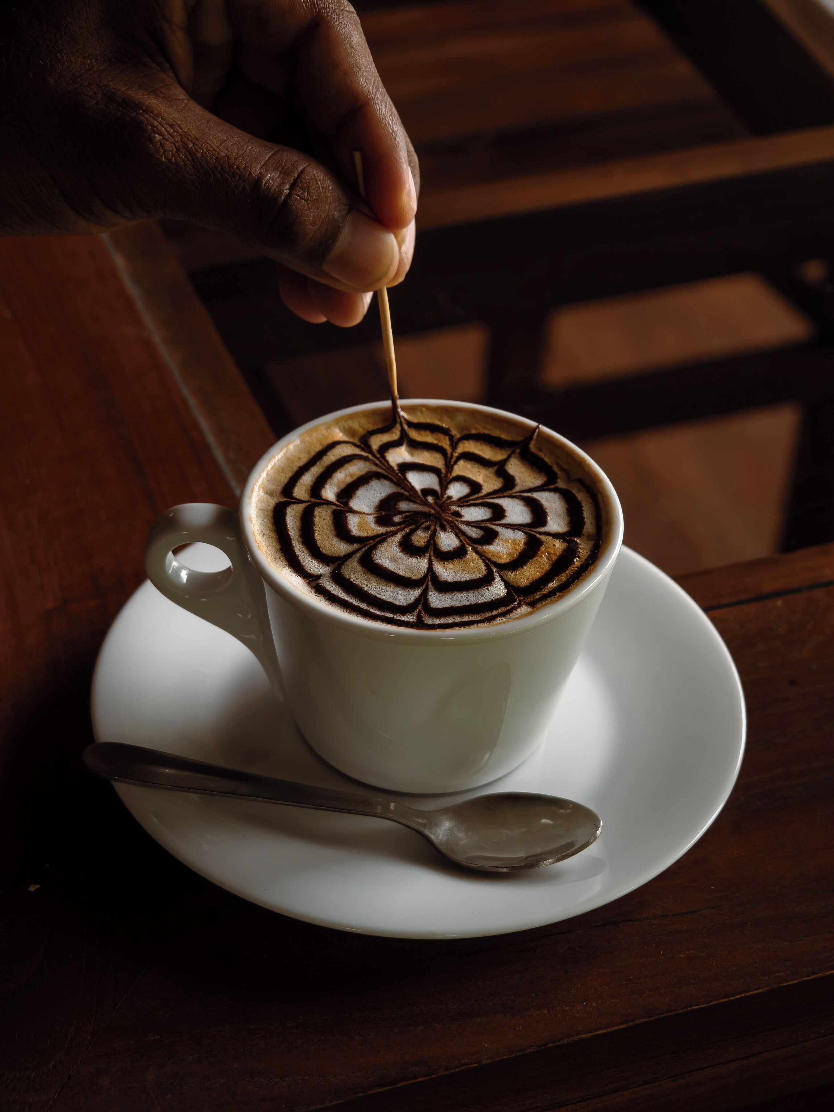 A hand holding a spoon, stirring a cup of coffee with a detailed latte art design.