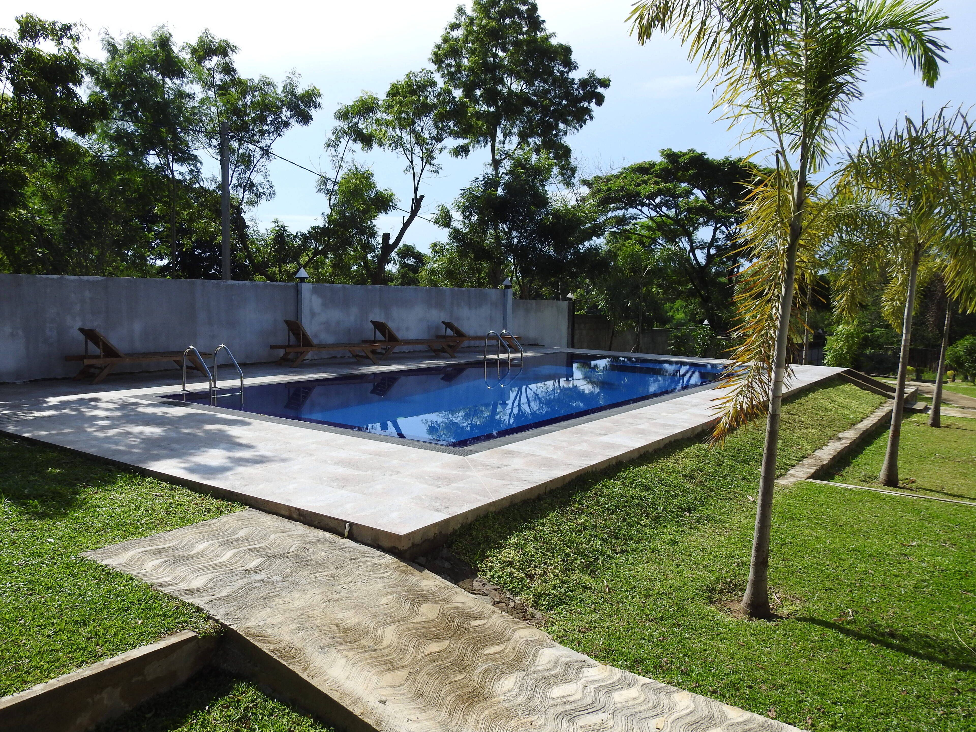 Una piscina rectangular con superficie de agua azul cristalina, rodeada por un patio de concreto con sillas reclinables de madera y escaleras metálicas.
