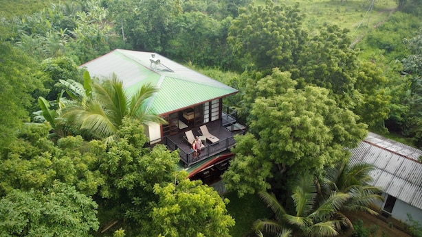 Une maison entourée d’une forêt dense, dotée d’un balcon et d’un espace extérieur aménagé pour s’asseoir.