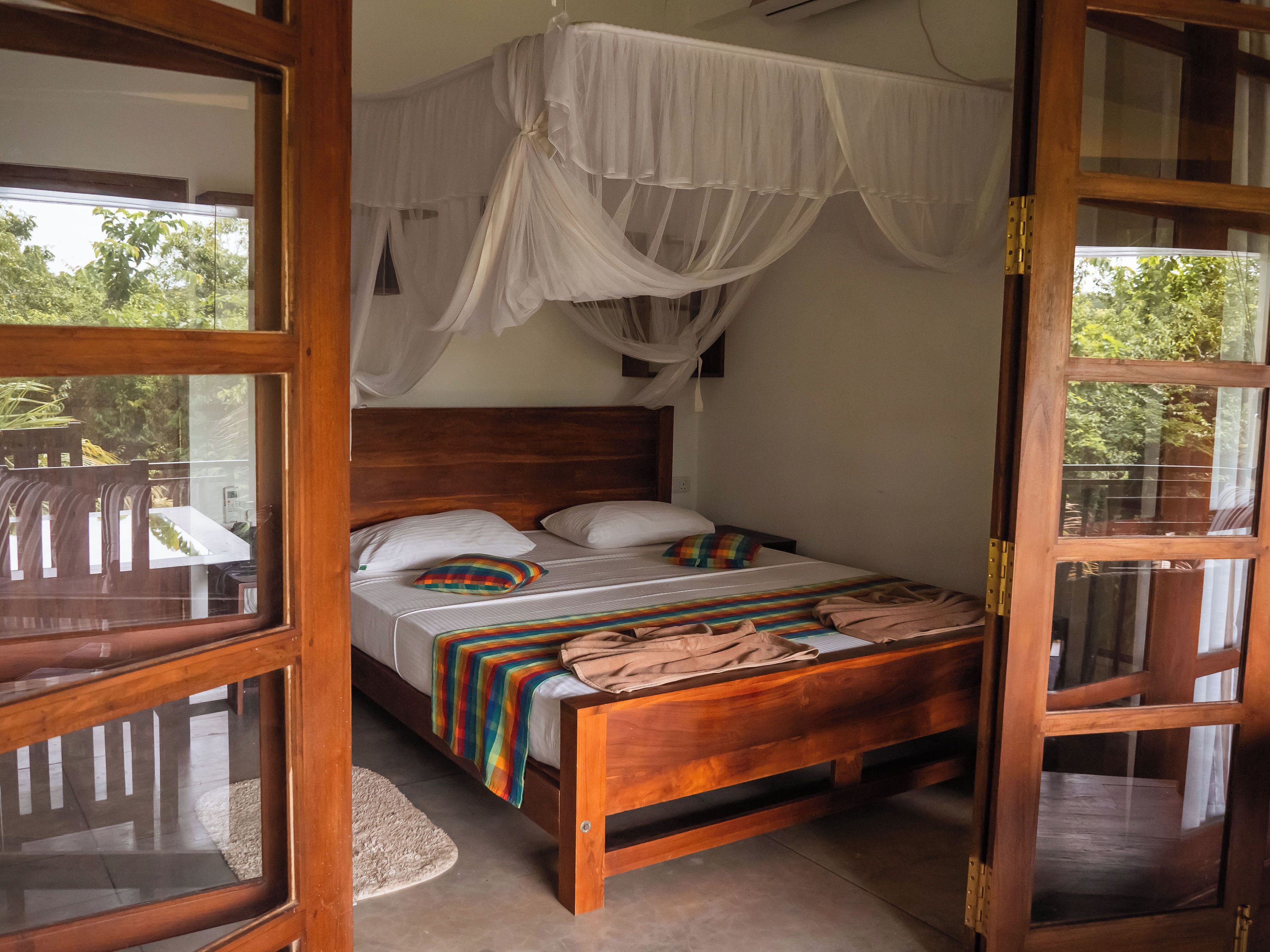 A room with a wooden bed, white curtains, and a view of greenery outside.