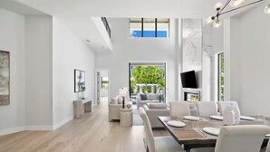 Une pièce spacieuse comprenant un coin repas, un ensemble de canapés, un téléviseur fixé au mur et une grande fenêtre donnant sur un paysage verdoyant.