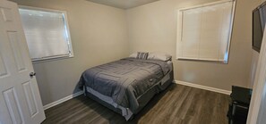 A bedroom with a bed, two windows with blinds, a door, and a small table.