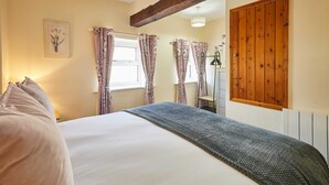 A bedroom with a bed, a window with curtains, a radiator, and a wooden door. - Host Stay Dairy Cottages (Ripon)