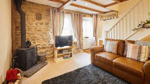 A cozy living room with a stone wall, a fireplace, a leather sofa, and a television on a wooden stand. - Host Stay Dairy Cottages (Ripon)