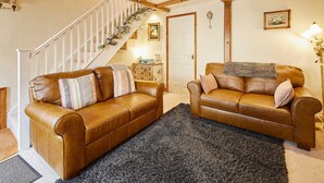 A living room with two brown leather sofas, a dark rug, a staircase with wooden beams, and a door with a framed picture above it. - Host Stay Dairy Cottages (Ripon)