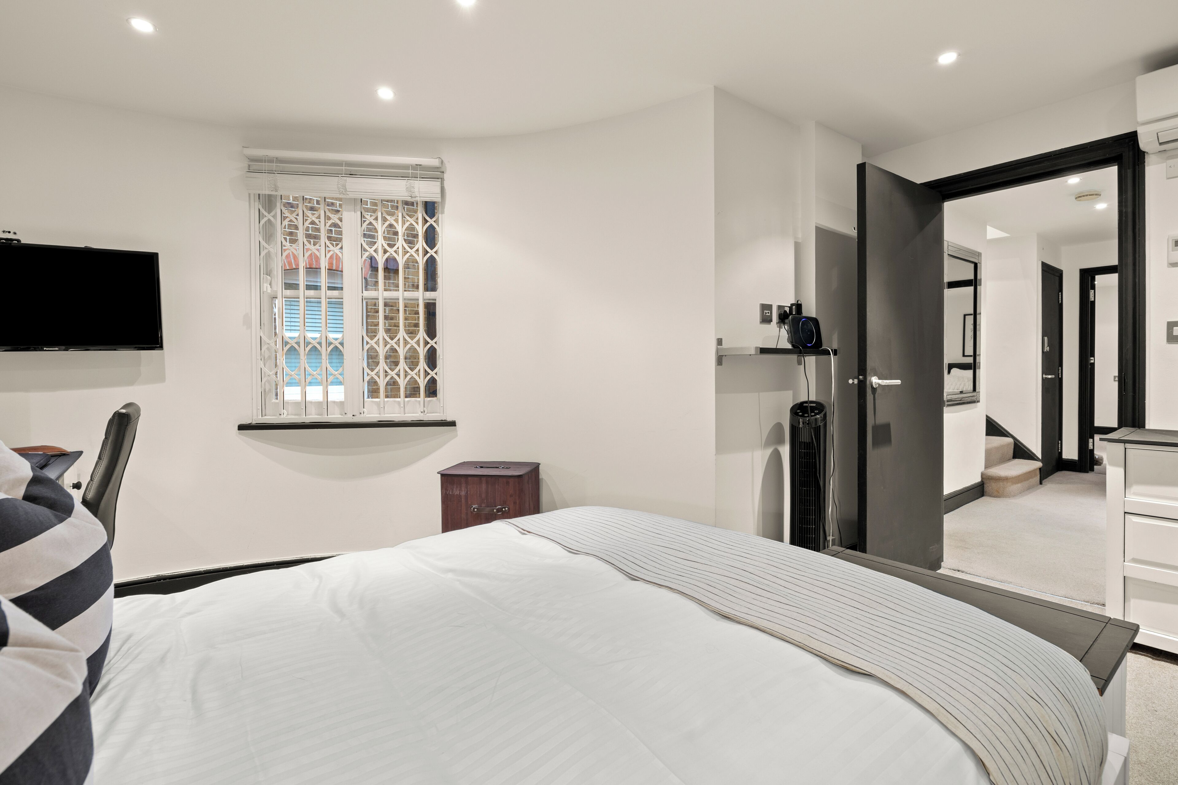 Quarto de hotel moderno com uma cama grande, televisão na parede, um pequeno armário de madeira, janela com grade decorativa e um espelho refletindo o quarto.