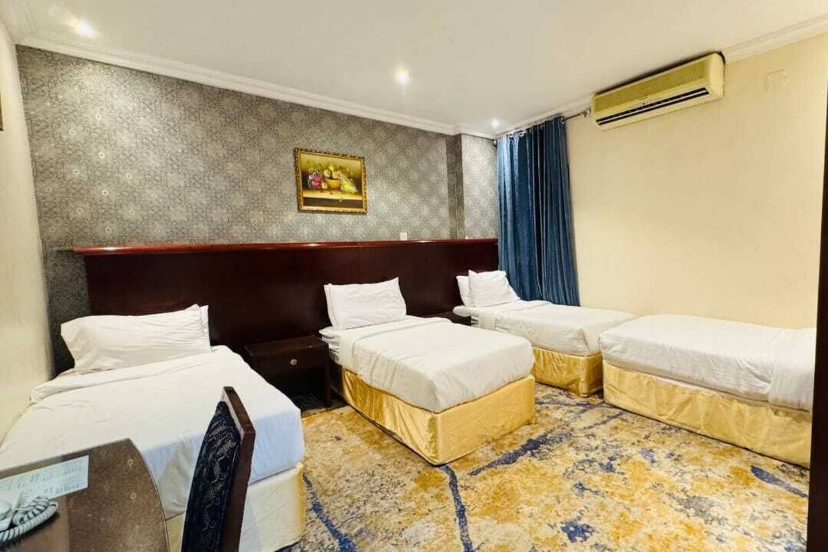 Ein Hotelzimmer mit drei Betten, einem Holz-Kopfteil, einem gemusterten Teppich und einer Klimaanlage.