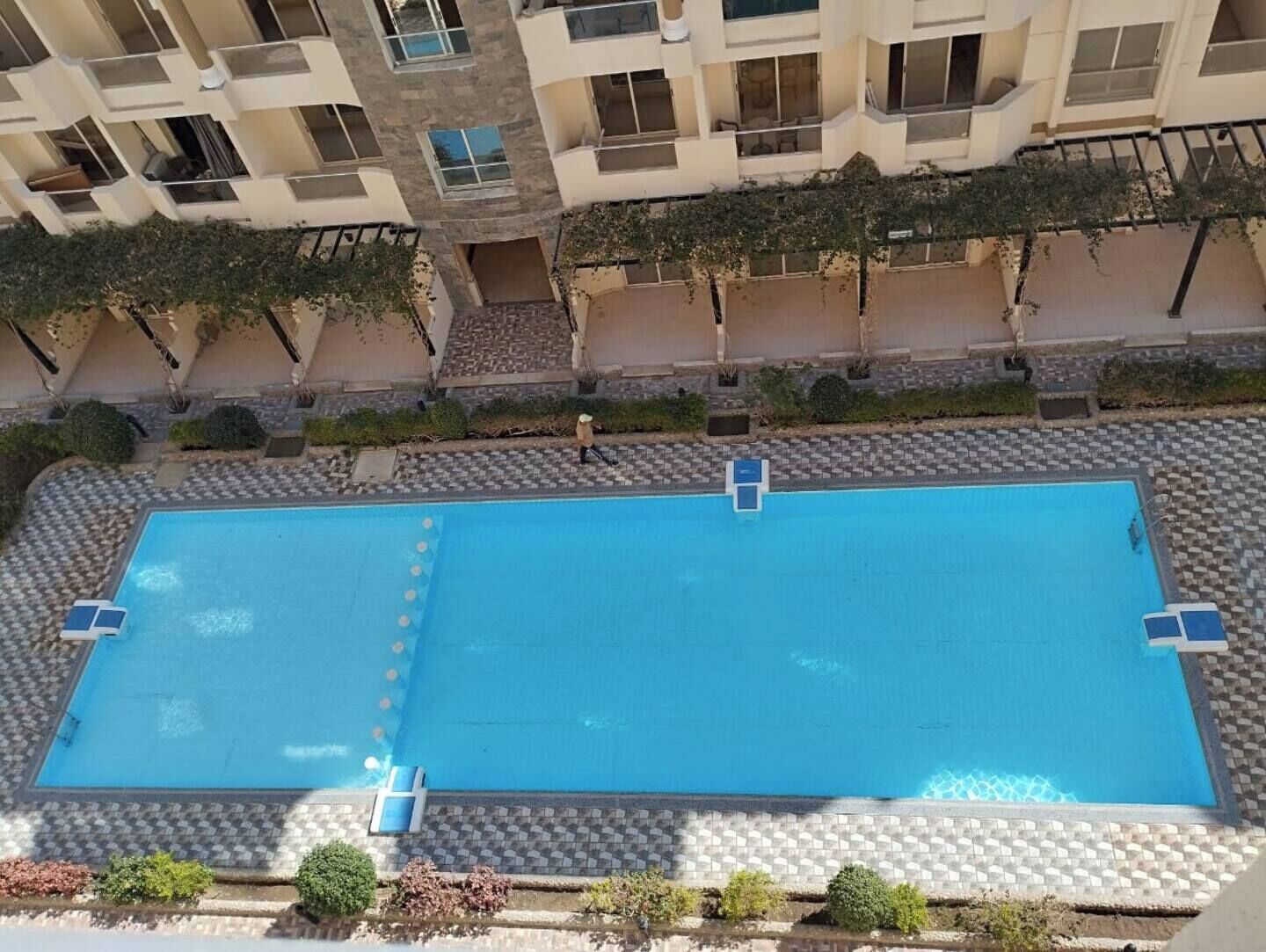 Une piscine rectangulaire entourée d’une terrasse carrelée, située dans un immeuble résidentiel à plusieurs étages doté de balcons.