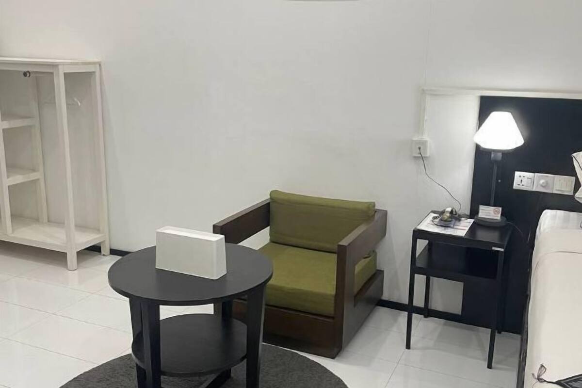 A room with a green chair, a small table with a box, a desk with a lamp, and a white wardrobe.
