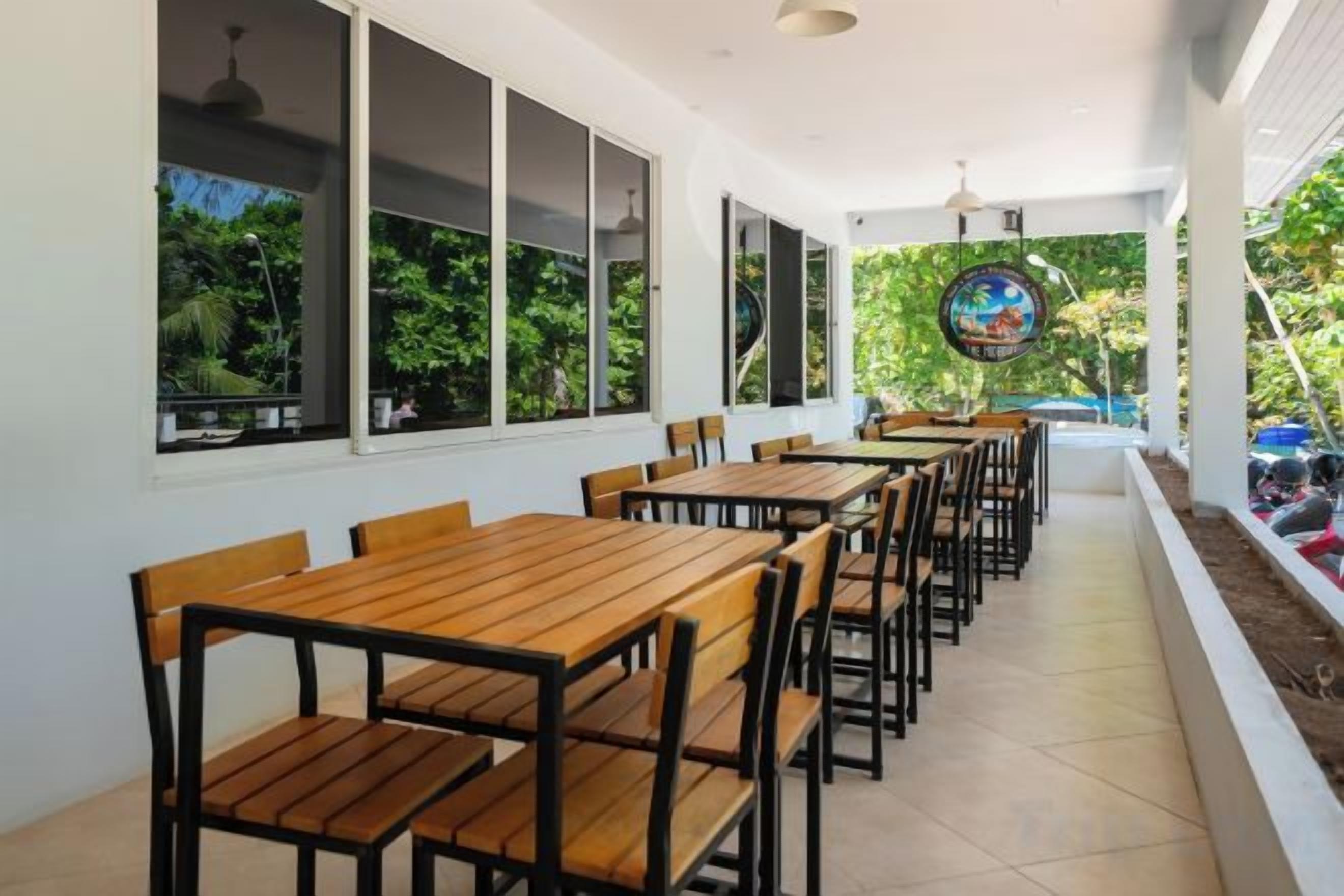 A covered outdoor seating area with wooden tables and chairs, large windows, and a view of greenery.