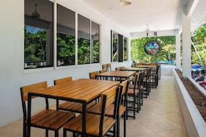 A covered outdoor seating area with wooden tables and chairs, large windows, and a view of greenery. - The Hideout Beachfront (Phuket)
