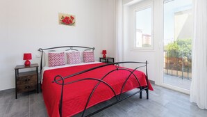 Ein Schlafzimmer mit roter Bettdecke, schwarzem Metallgestell, einem kleinen Nachttisch mit roter Lampe und einem Fenster mit Blick auf Gebäude und Grünflächen.