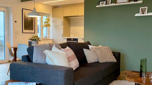 Un salon moderne avec un canapé gris, une table basse et un tapis blanc. On peut apercevoir une cuisine à travers une porte ouverte, ainsi qu’un coin repas avec une chaise et une table.