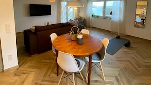 Un salon moderne avec un parquet en bois disposé en chevrons, une table à manger ronde en bois entourée de chaises blanches, un canapé marron foncé, un téléviseur à écran plat fixé au mur et une grande fenêtre offrant une belle vue.