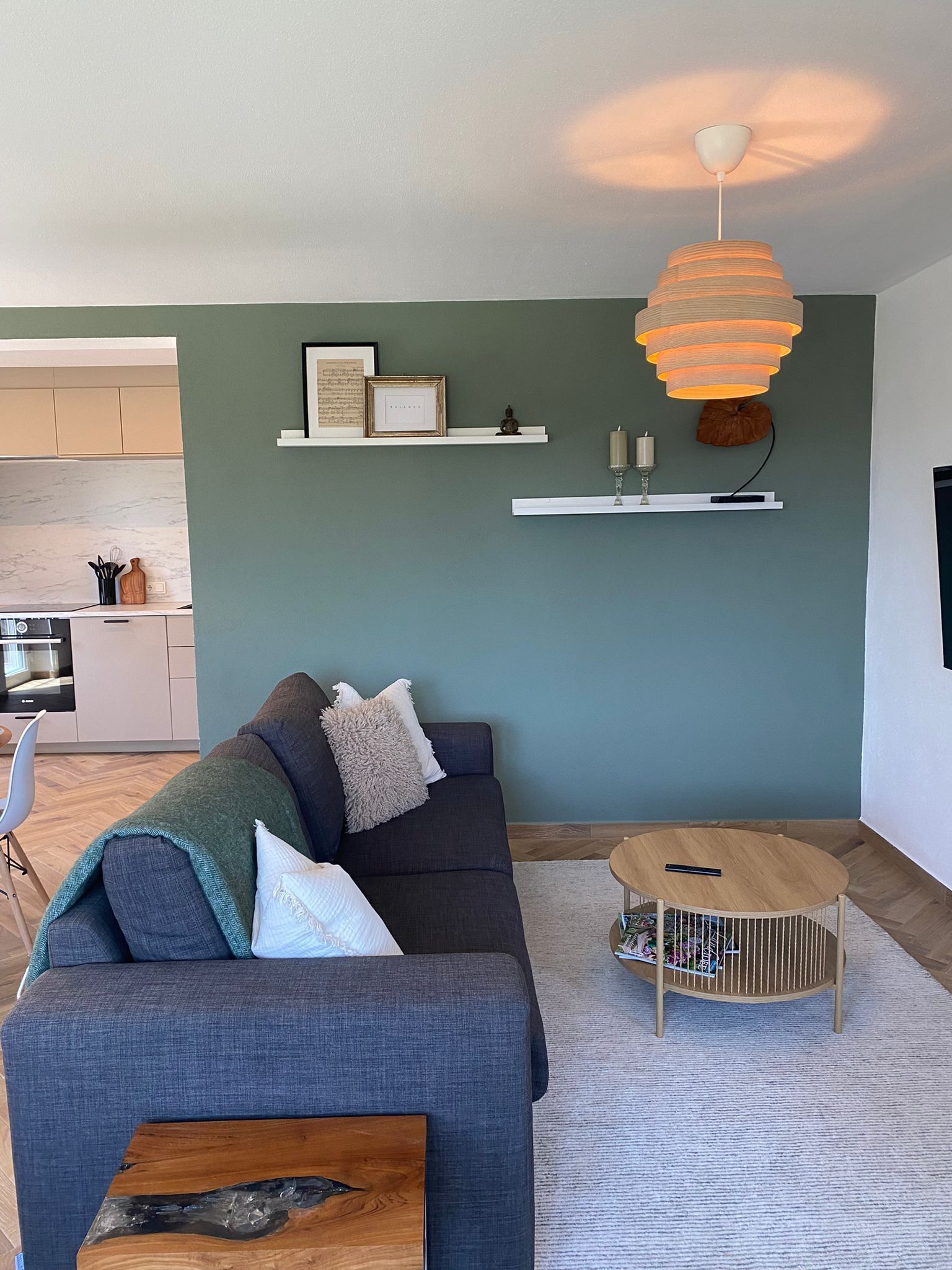 Een moderne woonkamer met een donkergrijze bank, een houten salontafel en een ronde bijzettafel met tijdschriften. Twee witte planken bevatten decoratieve objecten en er hangt een wandlamp.