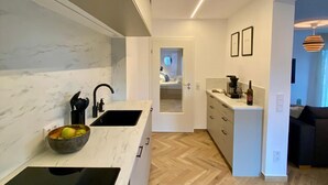 Une cuisine moderne avec un plan de travail en marbre, un évier noir et un saladier rempli de fruits. Le sol est en bois disposé en motif à chevrons, et on aperçoit le salon.