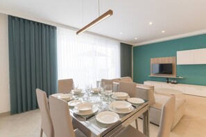 A modern dining area with a glass table set for four, surrounded by beige chairs. A flat-screen TV is mounted on the teal-colored wall, and a small kitchen area is visible in the background. - Property with Pool access - Xaghra near Marsalforn (Xagħra)
