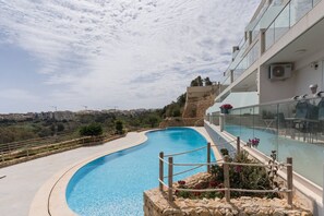 A curved swimming pool with a clear view of the surrounding landscape, a stone wall, and a modern building with glass railings. - Property with Pool access - Xaghra near Marsalforn (Xagħra)