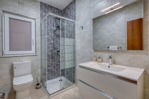 A modern bathroom with a glass-enclosed shower, a toilet, a sink with a mirror, and a wooden cabinet. - Apartment in Marsalforn – Beaches & Seaside Dining (Żebbuġ)