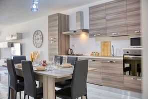 A modern kitchen with wooden cabinets, a dining table set for four, and a large clock on the wall. - Apartment in Marsalforn – Beaches & Seaside Dining (Żebbuġ)
