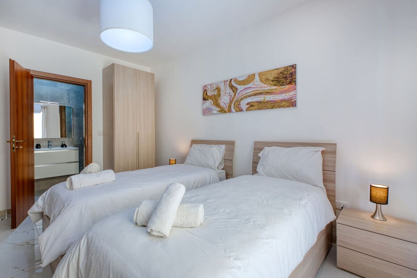 A hotel room with two beds, a wooden wardrobe, a bathroom with a mirror and sink, and a painting on the wall.
