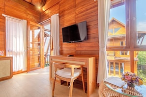 L’intérieur d’un chalet en bois est aménagé avec un téléviseur fixé au mur, un bureau en bois, une chaise en osier et une table en verre sur laquelle est posé un vase de fleurs.