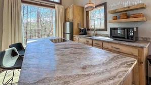 Una cocina con una isla grande, armarios de madera, un microondas y vistas a árboles desde las ventanas.