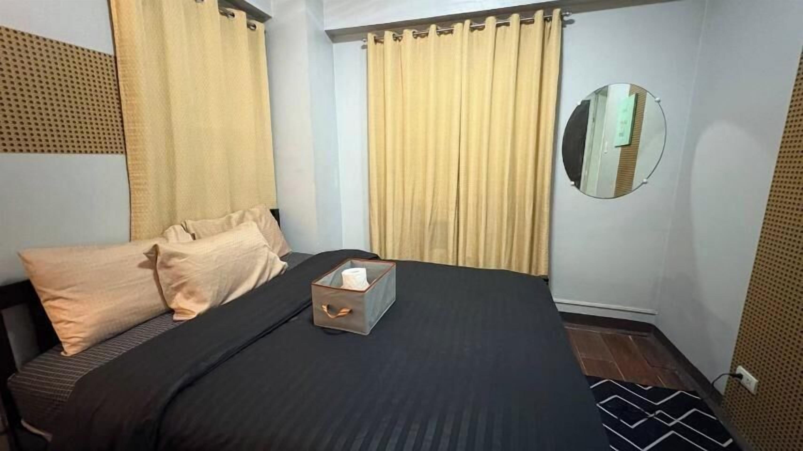 A bedroom with a bed, beige curtains, a mirror, and a bedside table with a tissue box.