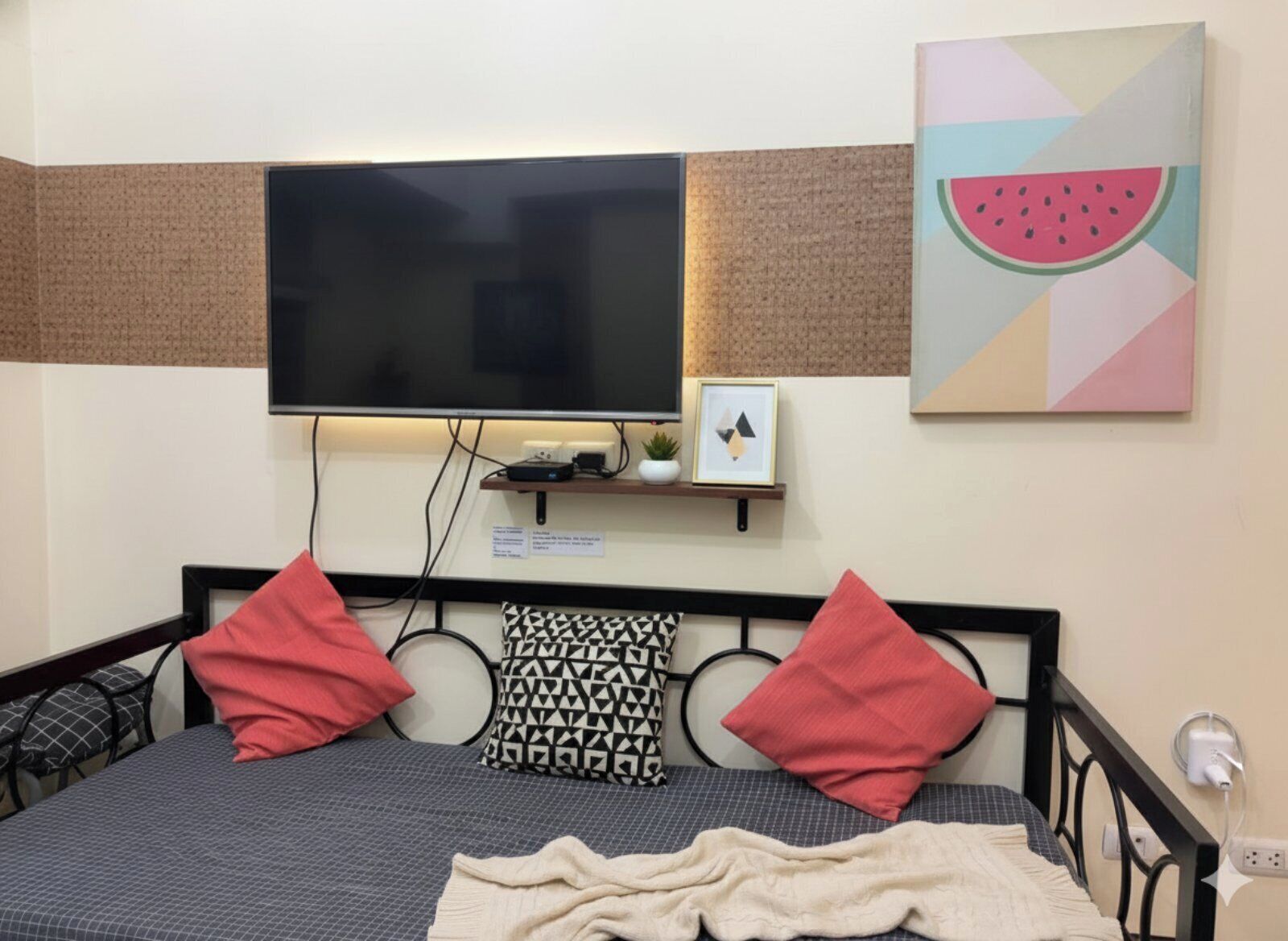 A neatly arranged hotel room with a bed, pillows, a flat-screen TV mounted on the wall, a small shelf with a plant and picture, and a wall art of a watermelon slice.