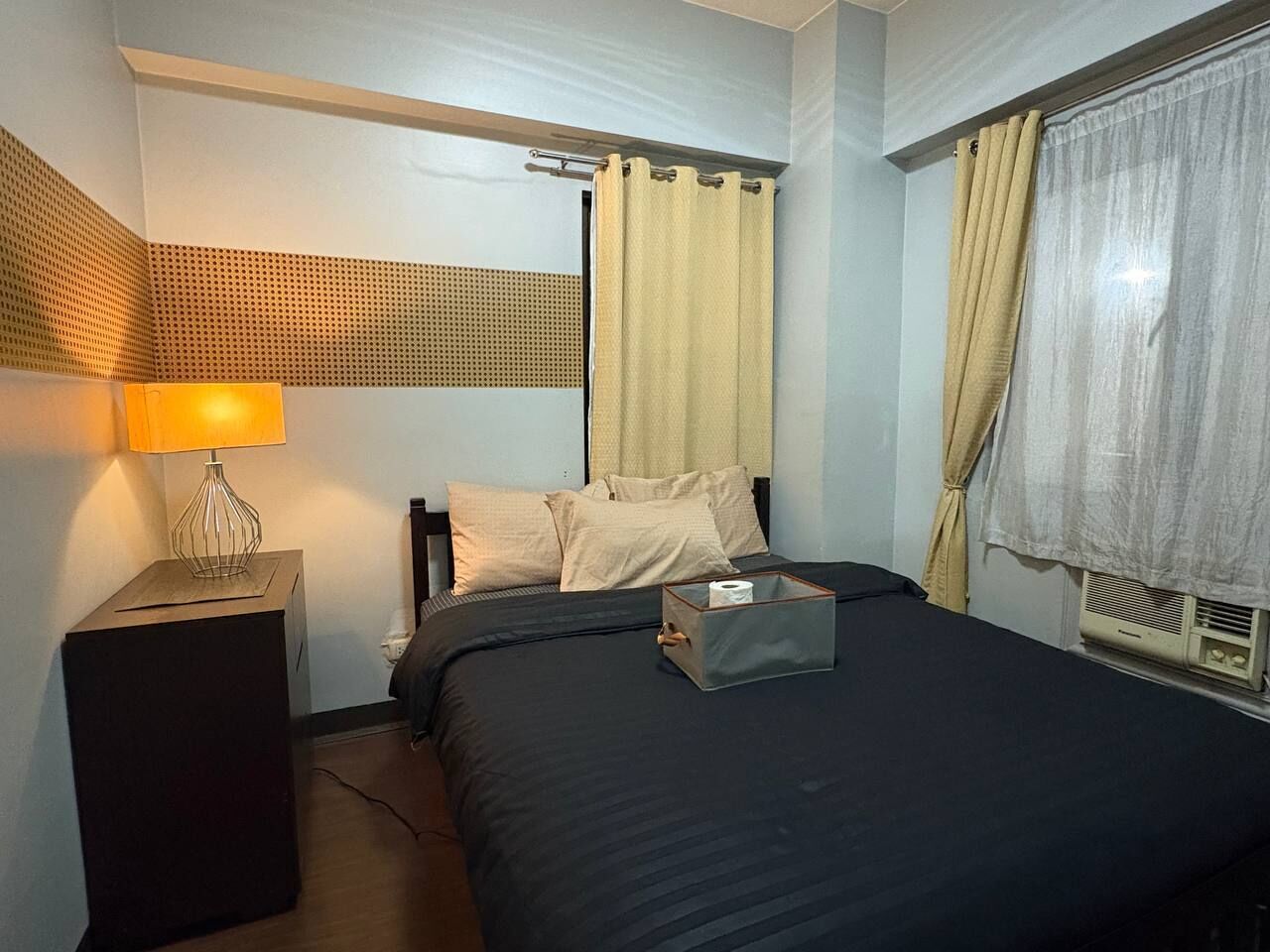 A hotel room with a bed, a bedside table with a lamp, a window with curtains, and a radiator.
