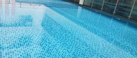 A large indoor swimming pool with clear blue water, surrounded by a modern building with large windows and a tiled floor.