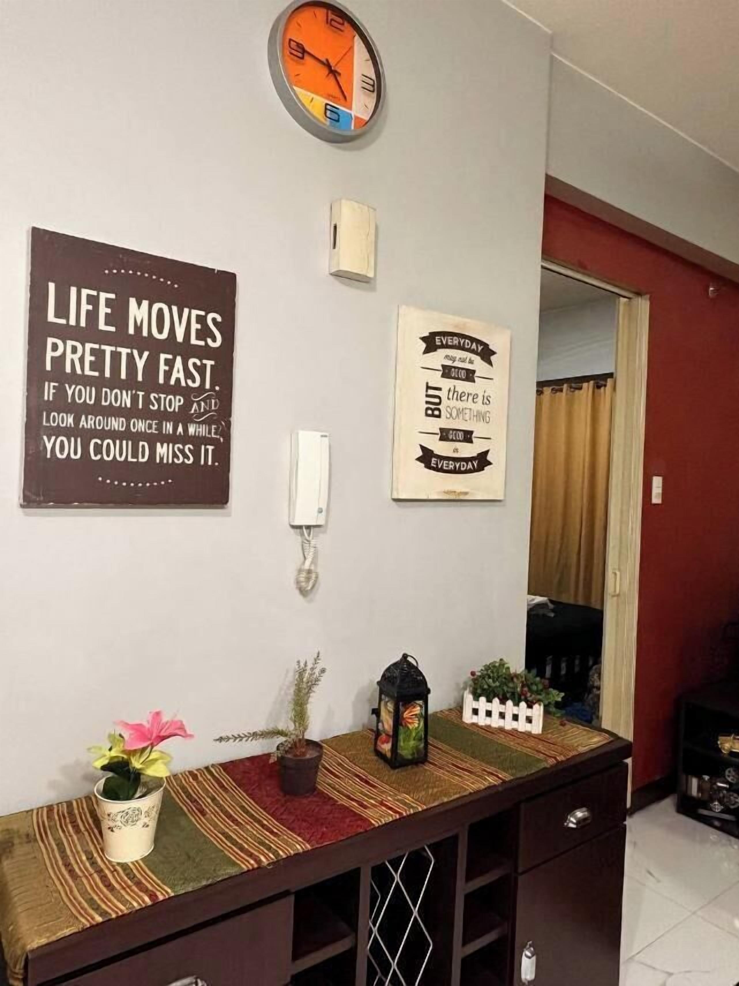 Una habitación con un reloj, dos carteles motivacionales, una mesita con una planta y un farol decorativo.