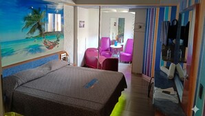 A hotel room with a bed, a mural of a beach scene, a small table with chairs, and a bathroom with a mirror and towel rack. - Hotel Puerto Angel (Nezahualcóyotl)