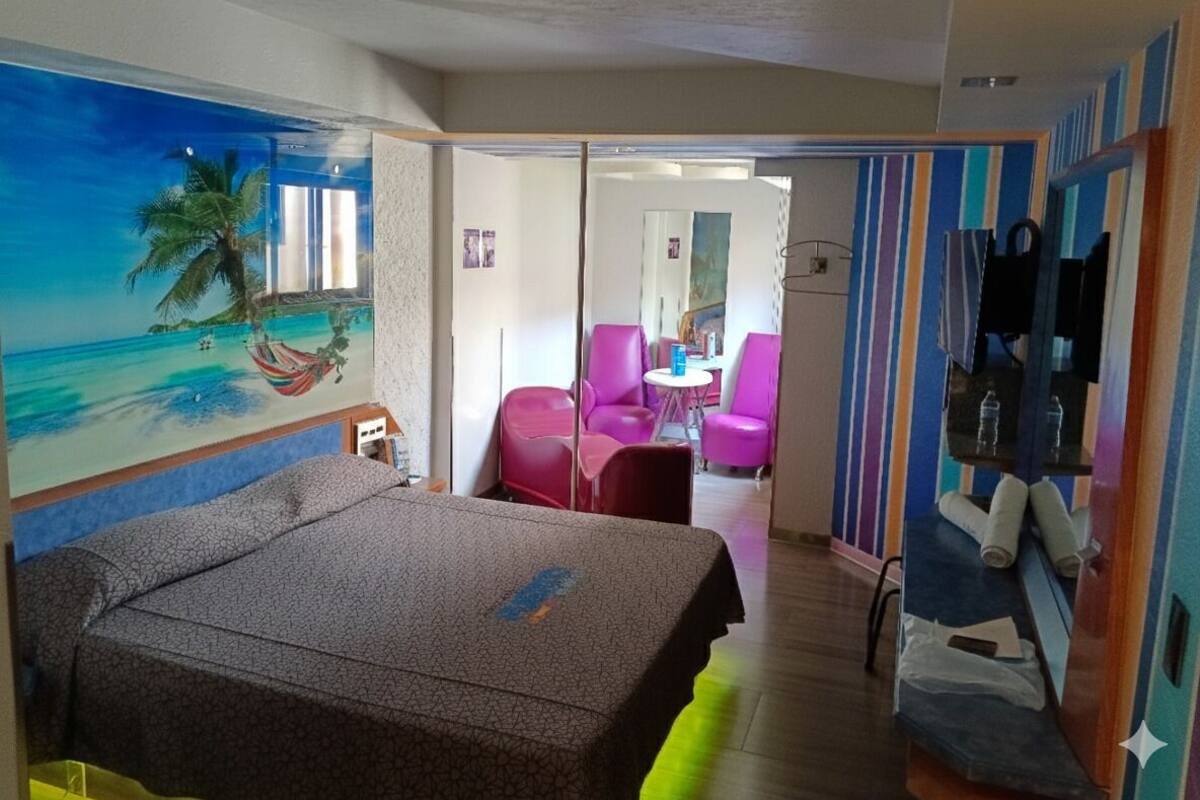 Habitación de hotel con cama, un mural de una playa, una mesita con sillas y un baño con espejo y toallero.