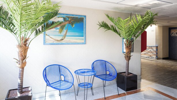 A lobby with three blue chairs, two potted palm trees, and a beach-themed painting on the wall. - Hotel Puerto Angel (Nezahualcóyotl)