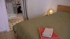 A bedroom with a bed, a nightstand with a lamp, and a bathroom visible through a curtain. - To complete (Bagnères-de-Bigorre)