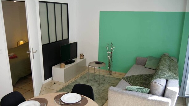 A modern living room with a green accent wall, a television on a white stand, a sofa, and a dining area with a table set for two. - To complete (Bagnères-de-Bigorre)