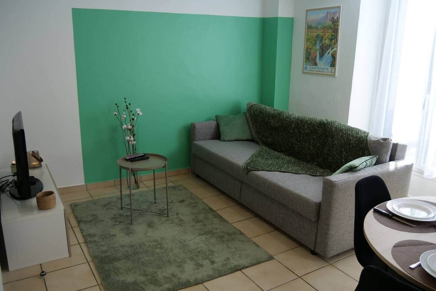 A living room with a green accent wall, a grey sofa, a small table with a remote control, and a dining area with a table set for two.