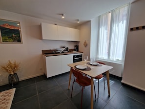 A compact kitchen with white cabinets, a dining table with chairs, and a window with sheer curtains.