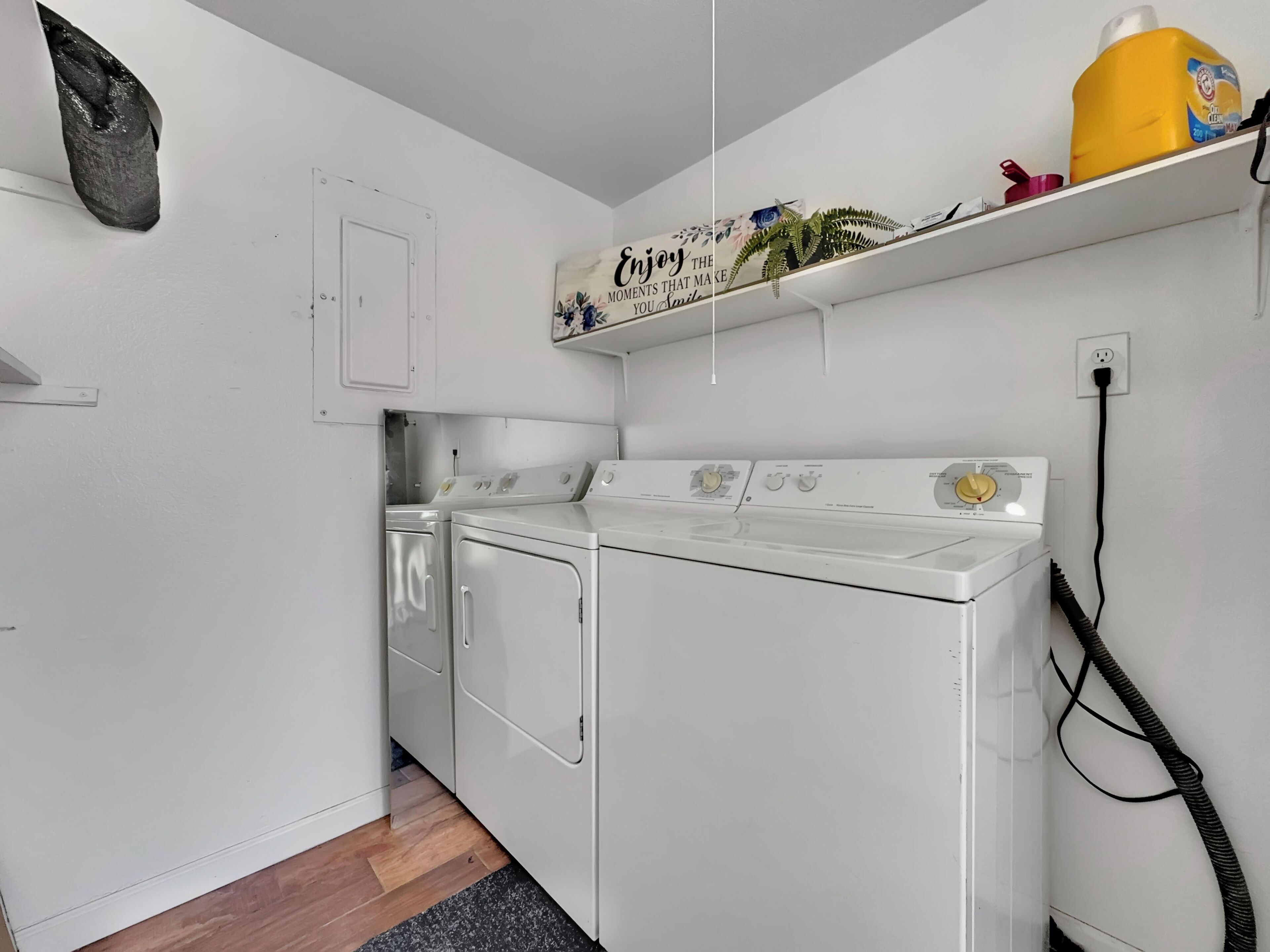 A laundry room with two white dryers, a shelf with a decorative sign, and a power outlet.