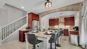A modern kitchen with a central island, bar stools, stainless steel appliances, and a staircase with a railing. - Puesta Del Sol (Queen Creek)