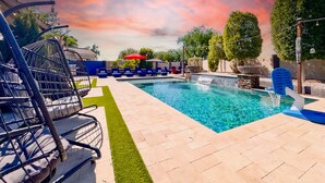 A swimming pool with a clear blue water feature, surrounded by lounge chairs and a red umbrella. - Puesta Del Sol (Queen Creek)