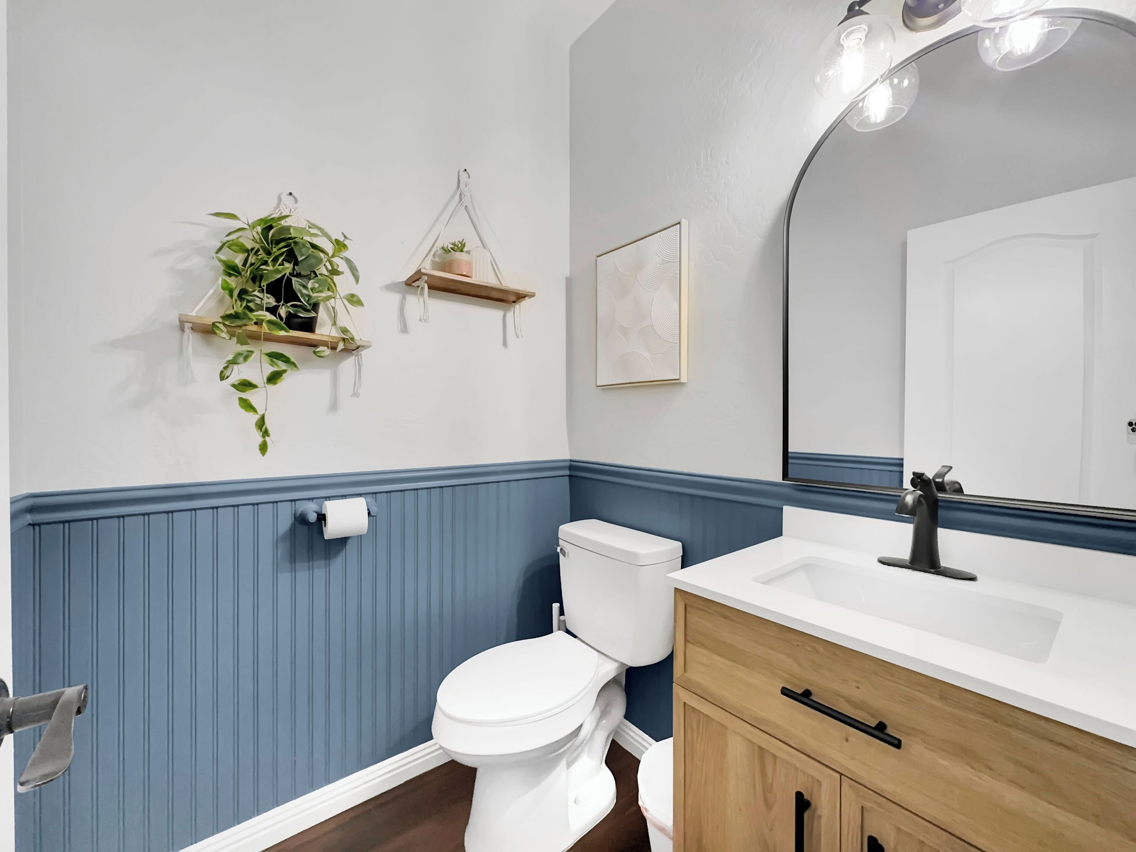 Un bagno con un water bianco, un mobile vanitario in legno e uno specchio. Sono presenti due mensole a parete con piante e un quadro incorniciato.