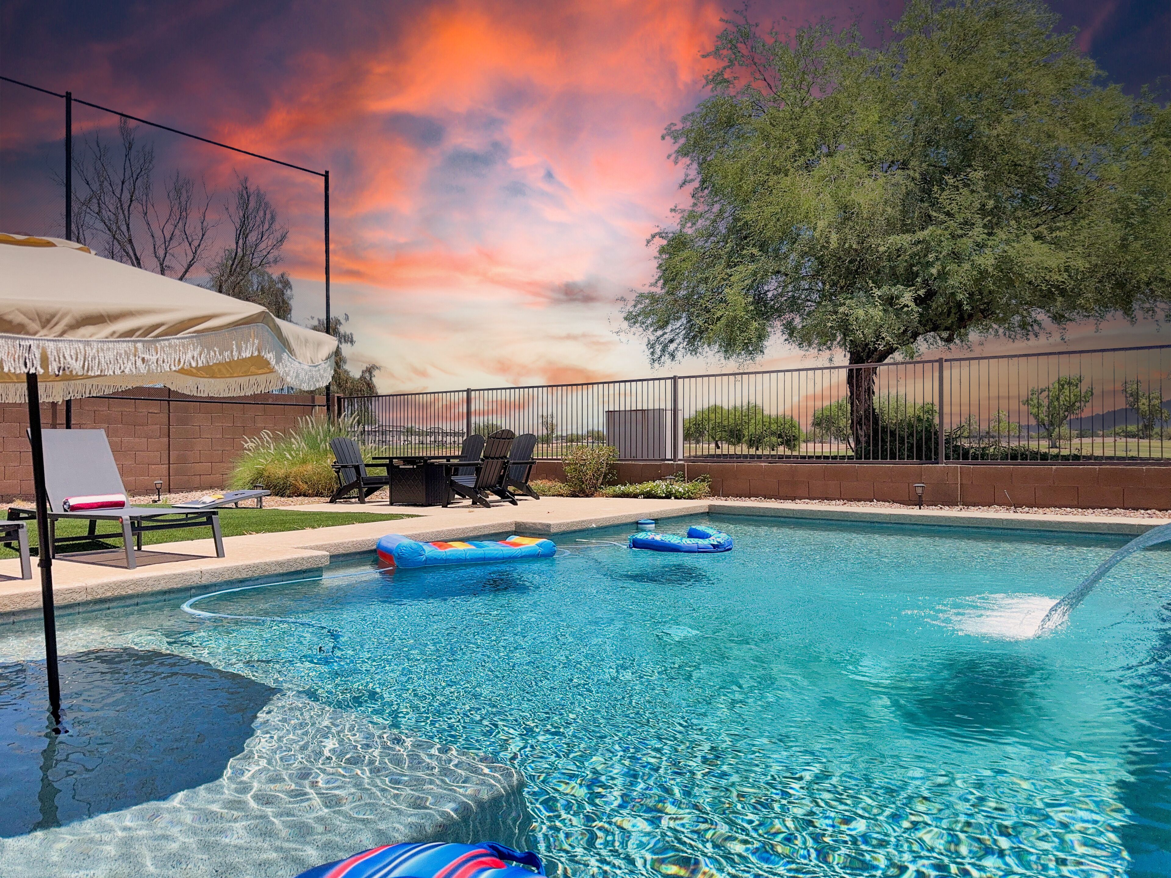 A swimming pool with clear water, surrounded by a fence, and a table with chairs under an umbrella.
